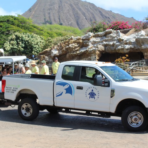 White truck and blue tram in tourist area with rocky backdrop.