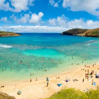 Panoramic view of a crowded beach with turquoise water and a rocky landscape under a blue sky.